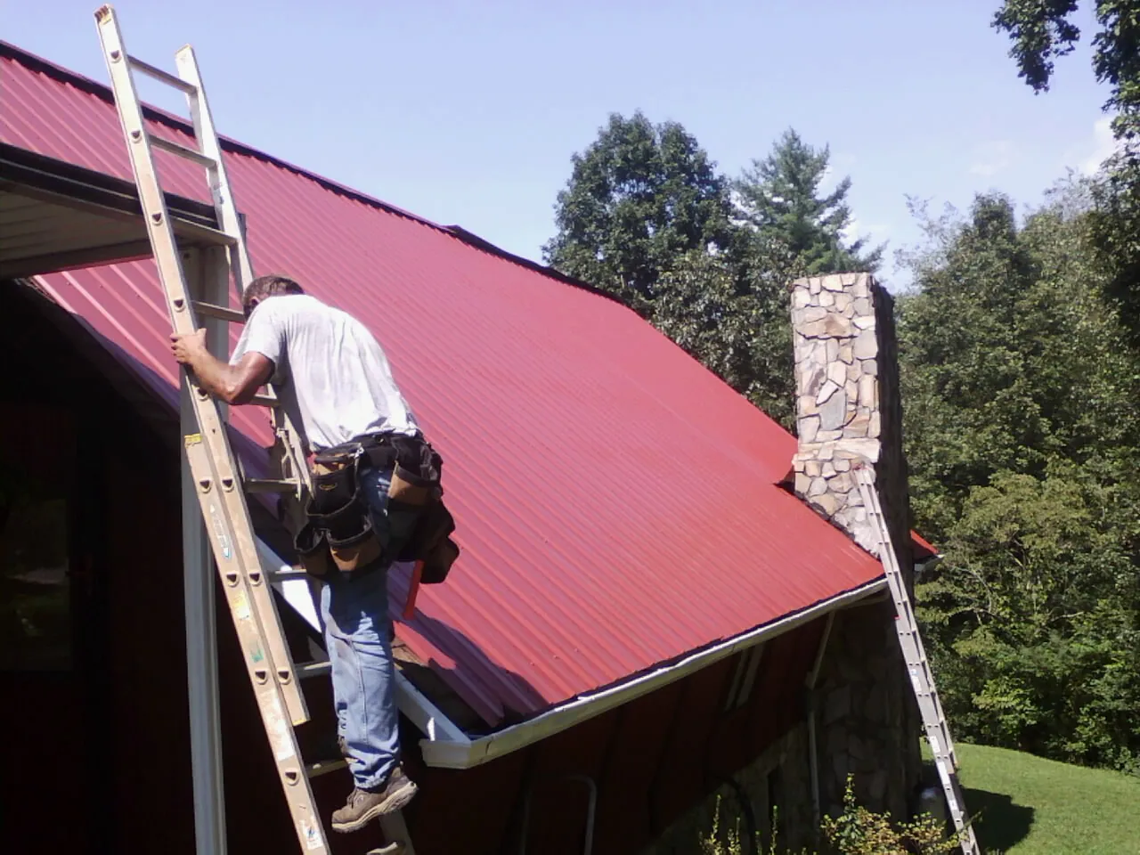 Roofer on a ladder beside a red metal roof and stone chimney — Soffit & Fascia Repair by Roofing Brothers in Waldron